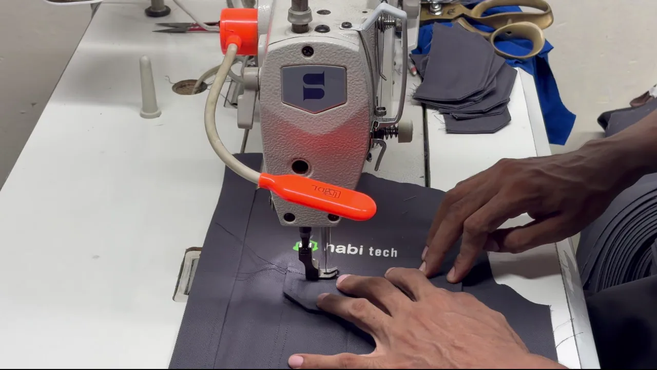 A KBwear machinist topstitching a charcoal habi tech work shirt panel with the embroidered chest logo already applied, filmed on the stitching floor of the KBwear factory in Lahore, Pakistan.