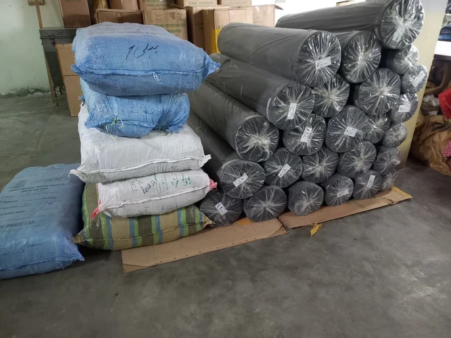 Charcoal fabric rolls, wrapped in plastic and tagged, stacked in the fabric store at the KBwear uniform factory in Lahore, Pakistan