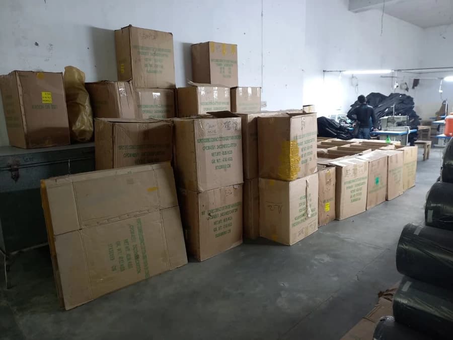 Packed and labelled export cartons stacked for dispatch at the KBwear uniform factory in Lahore, with a machinist working at the stitching line behind